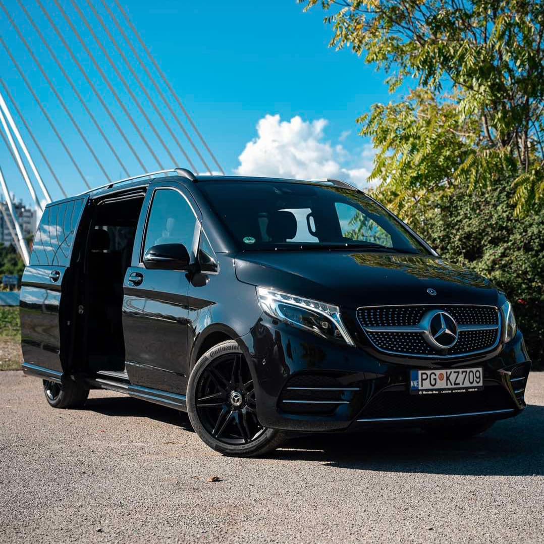 Black Mercedes-Benz V-Class van parked with its side door open, showcasing its sleek design and luxurious appearance, near Milenium bridge and clear blue sky in the background.